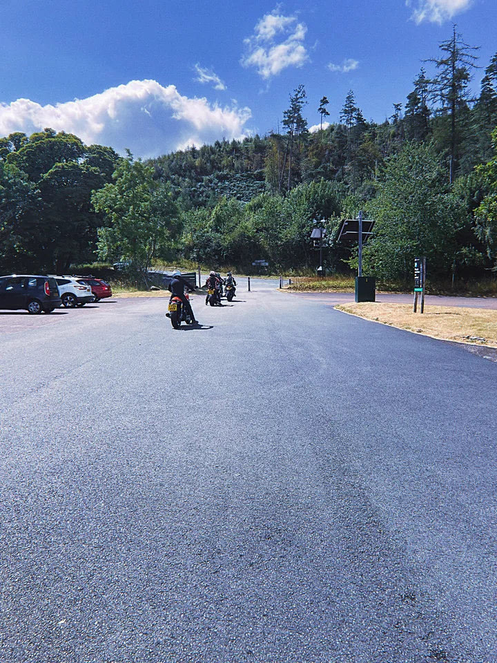 Motorcyclists leaving a car park beside a wooded hillside under a blue sky